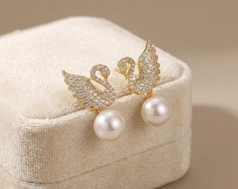 May include: A pair of gold-toned swan earrings. Each earring features a swan design encrusted with small, sparkling crystals and a large, round, white pearl. The earrings are displayed on a beige velvet surface.