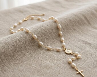 May include: A gold-toned rosary necklace with oval-shaped pearls. The necklace features a cross pendant and a medallion with a religious figure. The rosary is displayed on a neutral-colored fabric.