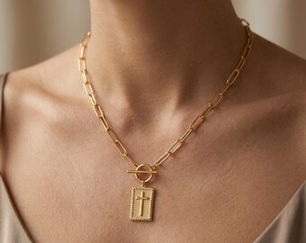 May include: Gold-toned necklace featuring a rectangular pendant with a cross design. The pendant is framed with a textured border and hangs from a chain with a toggle clasp. The necklace is worn around the neck.