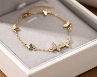 May include: A gold-colored bracelet featuring multiple butterfly charms. Some butterflies are solid, while others are adorned with small, sparkling stones. The bracelet is displayed in a white box with a gray interior, highlighting the jewelry's delicate design.