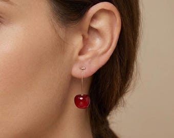 May include: A close-up of a person's ear wearing a cherry earring. The earring features a small, red cherry pendant suspended from a thin, gold-colored wire. The cherry has a glossy finish, and the earring post is also gold-colored.