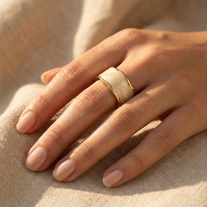 Puede incluir: Un anillo dorado con un diseño de esmalte crema veteado. El anillo tiene una banda ancha con un borde ondulado. El anillo está en un dedo y el fondo es de color neutro.