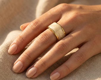 May include: A gold-toned ring with a cream-colored, marbled enamel design. The ring has a wide band with a wavy edge. The ring is on a finger, and the background is a neutral color.