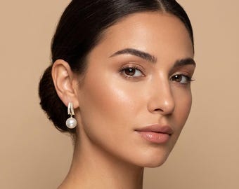 Puede incluir: Pendientes de aro dorados con una perla blanca colgante. El aro está incrustado con pequeños cristales transparentes. Los pendientes se presentan sobre un fondo neutro, destacando el elegante diseño de la joya. Un accesorio clásico.
