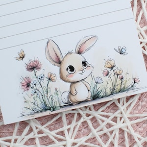 Könnte beinhalten: Ein weißer Notizblock mit horizontalen Linien und einer Aquarell-Illustration eines kleinen beigen Hasen, der zwischen rosa und gelben Blumen mit zwei Schmetterlingen steht. Die Zeichnung hat weiche Konturen und Pastellfarben auf weißem Grund.
