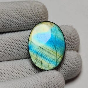 Puede incluir: Una piedra preciosa de labradorita de forma ovalada con una superficie pulida que presenta destellos iridiscentes de azul vibrante y amarillo pálido. La piedra muestra fracturas y estrías internas naturales en su cara lisa, redondeada y reflectante.