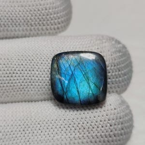 Può includere: Una pietra preziosa di labradorite di forma quadrata con angoli arrotondati, che mostra un vibrante riflesso iridescente blu e verde acqua sulla sua superficie lucida, con fratture interne visibili e toni grigio scuro in tutta la pietra.