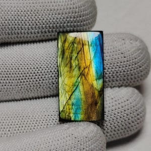 May include: A rectangular labradorite gemstone featuring a polished surface with vibrant flashes of iridescent blue and golden yellow colours, displaying natural internal fractures and a smooth, reflective finish across the entire stone face.