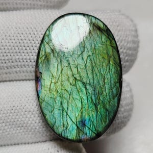 Può includere: Una pietra preziosa di labradorite di forma ovale con una superficie lucida e liscia caratterizzata da un vibrante gioco di iridescenza verde e blu. La pietra mostra una complessa rete di fratture interne e inclusioni naturali nella struttura.