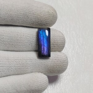 May include: A rectangular labradorite gemstone featuring a polished surface with a vibrant blue and purple iridescent flash. The stone displays natural internal fractures and a dark base color, showcasing a distinct color play across its face.