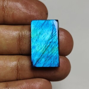 May include: A rectangular labradorite gemstone featuring a vibrant blue and green iridescent surface with visible internal striations and a polished finish. The stone displays a distinct play of colour across its flat, smooth, and reflective face.