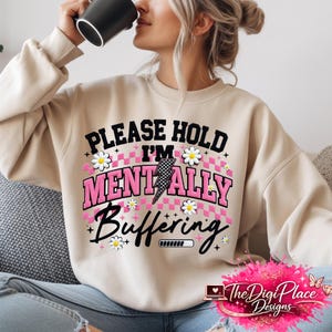 Peut inclure: Sweat-shirt beige avec l'inscription "PLEASE HOLD I'M MENTALLY BUFFERING" en noir et rose. Le motif comprend un éclair, des marguerites et une barre de chargement. Le logo The Digi Place Designs est en bas à droite.