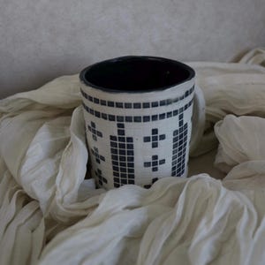 May include: A ceramic mug with a white base and a black interior. The exterior features a geometric pattern of black squares and crosses. The mug is set against a backdrop of draped, off-white fabric.