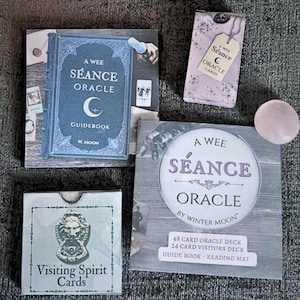 May include: A collection of A Wee Séance Oracle items including a blue guidebook, a small pink card box, a square box labeled Visiting Spirit Cards with a lion door knocker illustration, a larger square box set, and a smooth round pink stone.