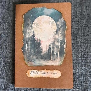 May include: A rectangular notebook with a brown kraft paper cover. The front features a centered, deckle-edged illustration of a full moon over a dark forest landscape. Below the illustration is a small, torn-edge label with the text Field Companion.