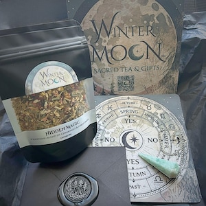 May include: A black package of "Hidden Magic" tea with a "Winter Moon" logo, a divination board, and a green crystal pendant. The tea bag is filled with dried herbs and flowers. The packaging includes a box and an envelope with a wax seal.