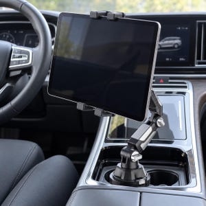 Könnte beinhalten: Ein schwarzer und silberner Tablethalter, der im Getränkehalter eines Fahrzeugs montiert ist. Das Gerät verfügt über einen verstellbaren Gelenkarm und eine Federklemme, die ein Tablet horizontal über der Mittelkonsole des Autos fixiert.