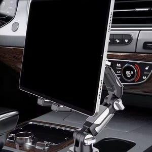 Peut inclure: Une tablette noire est fixée sur un support de téléphone de voiture argenté et noir. Le support est fixé au tableau de bord de la voiture. L'intérieur de la voiture présente des boiseries et un affichage numérique.