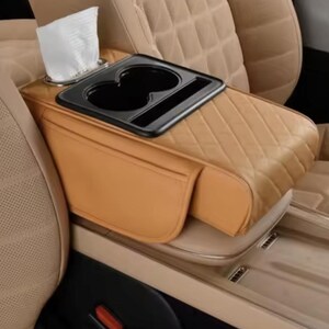 May include: Tan leather car console with a black drinks tray and tissue holder. The console features a quilted design and is positioned between two beige car seats. A small pocket is visible on the side.