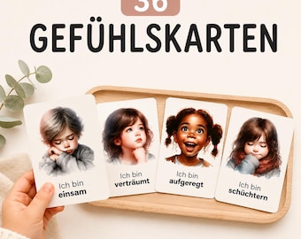 May include: A set of 36 feeling cards titled 36 GEFÜHLSKARTEN. Four cards are displayed, each featuring a child's illustration and German text: Ich bin einsam, Ich bin verträumt, Ich bin aufgeregt, and Ich bin schüchtern. The cards rest on a wooden tray.