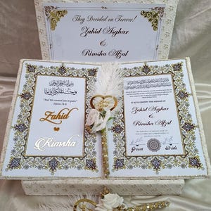 May include: A wedding keepsake set with a decorative box and book, adorned with gold detailing. The set includes a pen with a heart-shaped embellishment and white roses. The text on the box reads "They Decided on Forever!" and the names Zahid Asghar and Rimsha Afzal.