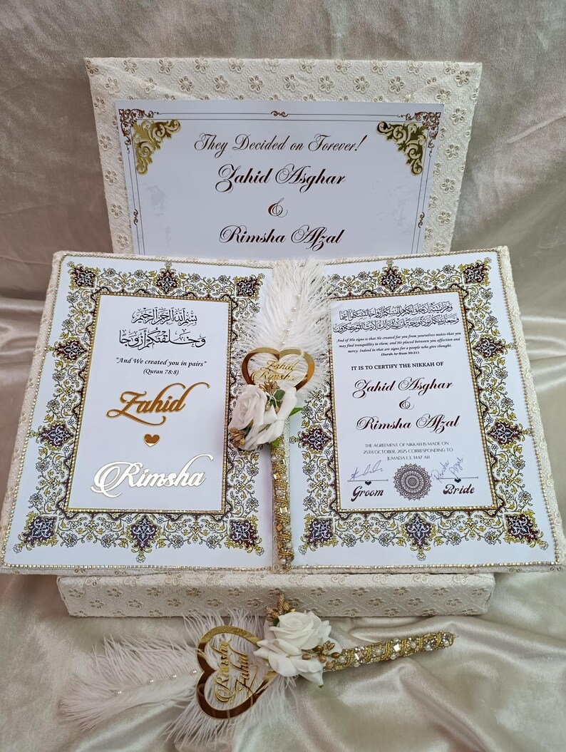 May include: A wedding keepsake set with a decorative box and book, adorned with gold detailing. The set includes a pen with a heart-shaped embellishment and white roses. The text on the box reads "They Decided on Forever!" and the names Zahid Asghar and Rimsha Afzal.