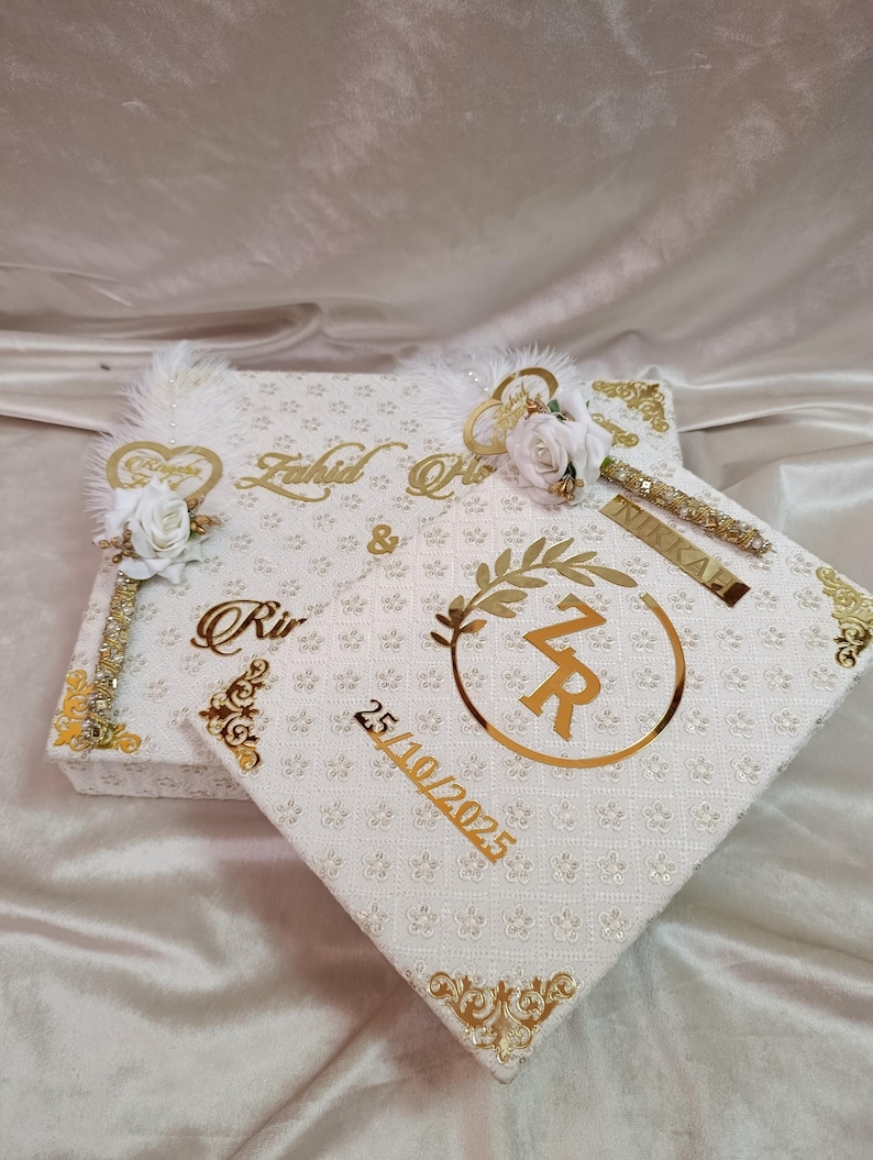 May include: White and gold decorative boxes with floral and feather details. The boxes have gold lettering and the date 25/10/2025. These boxes are likely for a wedding or special occasion.