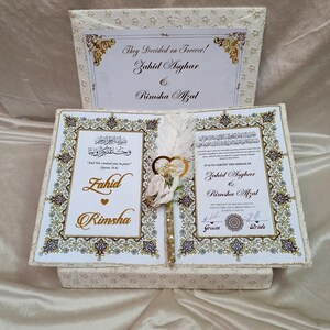 May include: A cream-coloured wedding box, open to reveal two pages with gold calligraphy and the names Zahid Asghar and Rimsha Afzal. The box has a patterned exterior and a decorative feather and flower arrangement in the centre.