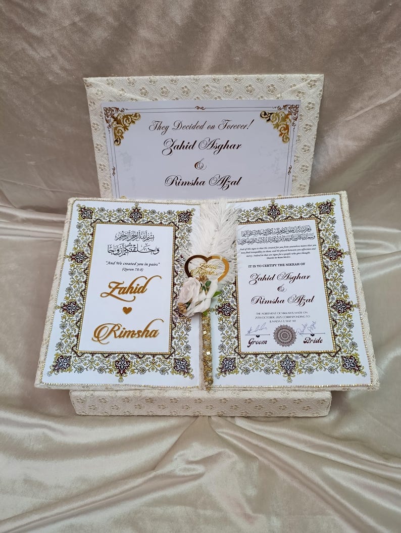 May include: A cream-coloured wedding box, open to reveal two pages with gold calligraphy and the names Zahid Asghar and Rimsha Afzal. The box has a patterned exterior and a decorative feather and flower arrangement in the centre.