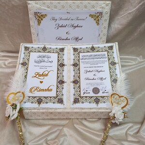May include: A cream wedding gift box with gold detailing and calligraphy. The box contains a decorative card with the names Zahid Asghar and Rimsha Afzal. Two decorative pens with gold accents and white feathers are included.