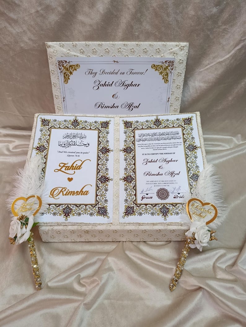 May include: A cream wedding gift box with gold detailing and calligraphy. The box contains a decorative card with the names Zahid Asghar and Rimsha Afzal. Two decorative pens with gold accents and white feathers are included.