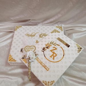 May include: Two white decorative boxes with gold detailing. The boxes feature a floral pattern and gold trim. One box is printed with the names "Fahid & Flo". The other box has the initials "ZR" and the date "25/10/2025". A decorative feather and flower are attached.