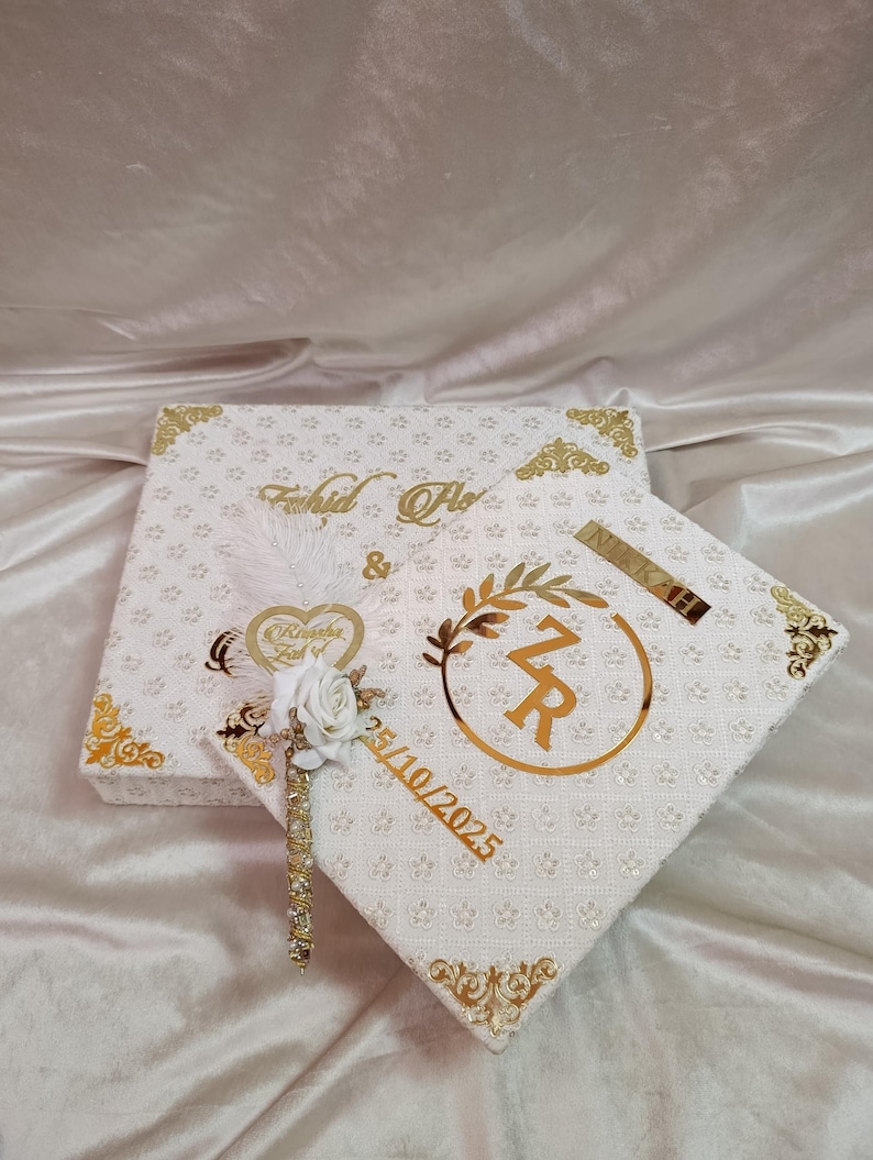 May include: Two white decorative boxes with gold detailing. The boxes feature a floral pattern and gold trim. One box is printed with the names "Fahid & Flo". The other box has the initials "ZR" and the date "25/10/2025". A decorative feather and flower are attached.