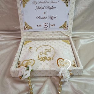 May include: A cream-coloured wedding box with gold detailing, featuring the text "They Decided on Forever!" and names. Inside, a decorated book and two pens adorned with white roses and gold embellishments are visible. The date "OCT 25 2025" is also present.