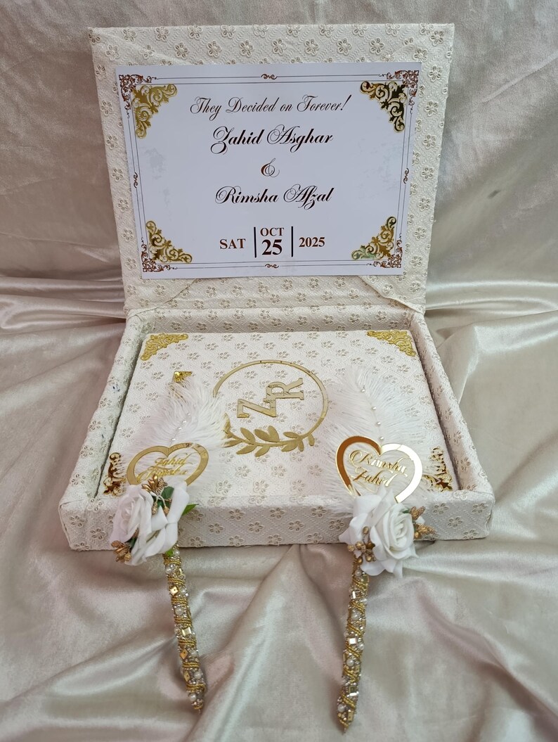 May include: A cream-coloured wedding box with gold detailing, featuring the text "They Decided on Forever!" and names. Inside, a decorated book and two pens adorned with white roses and gold embellishments are visible. The date "OCT 25 2025" is also present.