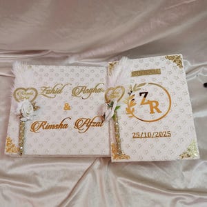 May include: Two white wedding guest books with gold detailing. The left book features the names "Fahid Asgha" and "Rimsha Afzal" in gold script. The right book has the word "Nikkah" and the date "25/10/2025".