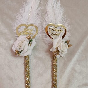 May include: Two decorative pens featuring white feathers, gold heart-shaped name tags, and white rose accents. The pen barrels are adorned with gold and clear crystals. The names "Farid Rimesha" and "Rimesha Zahid" are inscribed on the heart-shaped tags.