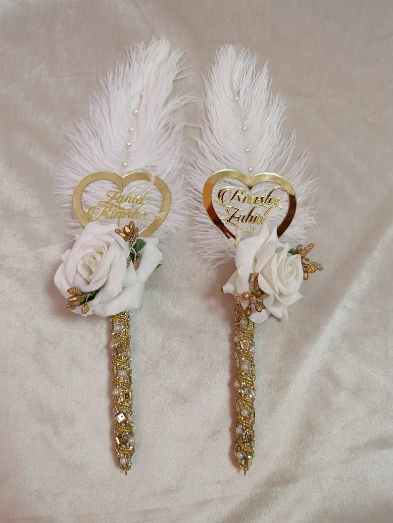 May include: Two decorative pens featuring white feathers, gold heart-shaped name tags, and white rose accents. The pen barrels are adorned with gold and clear crystals. The names "Farid Rimesha" and "Rimesha Zahid" are inscribed on the heart-shaped tags.