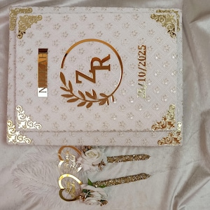May include: A white wedding guest book with gold detailing, displaying the initials "ZR" in a gold circle with a floral design. Includes two pens with gold decoration and white flowers. The date "15/10/2025" is also visible.