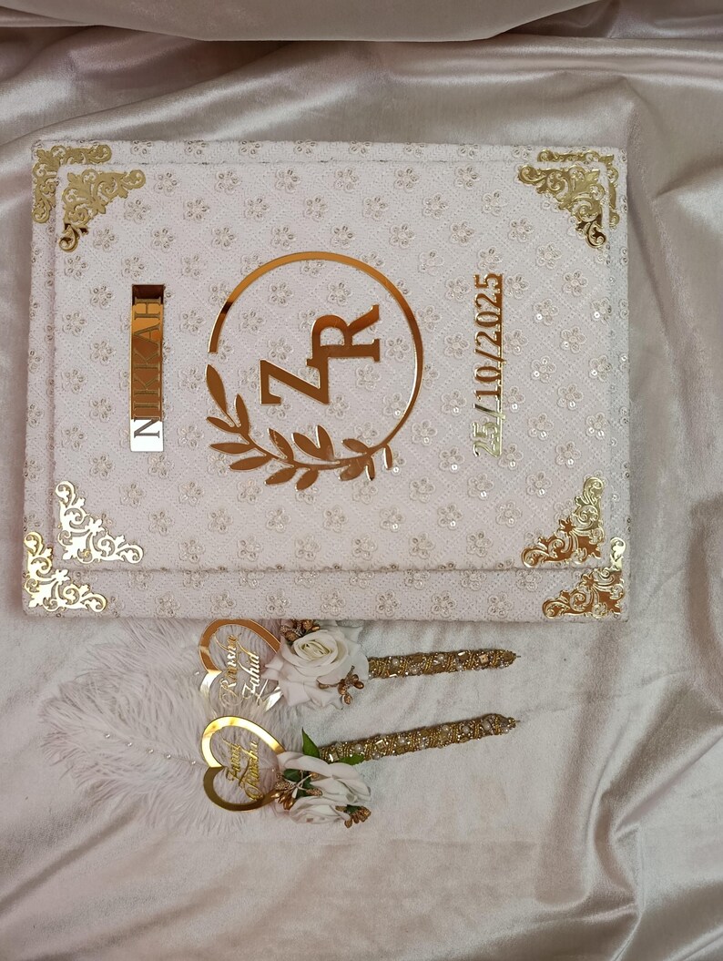 May include: A white wedding guest book with gold detailing, displaying the initials "ZR" in a gold circle with a floral design. Includes two pens with gold decoration and white flowers. The date "15/10/2025" is also visible.