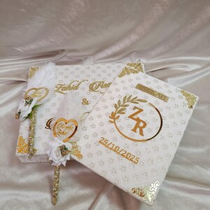 May include: Two white decorative boxes with gold detailing. One box features the names "Fahid & Asma" in gold script. The other box has the initials "ZR" within a gold wreath, with the date "25/10/2025" in gold.