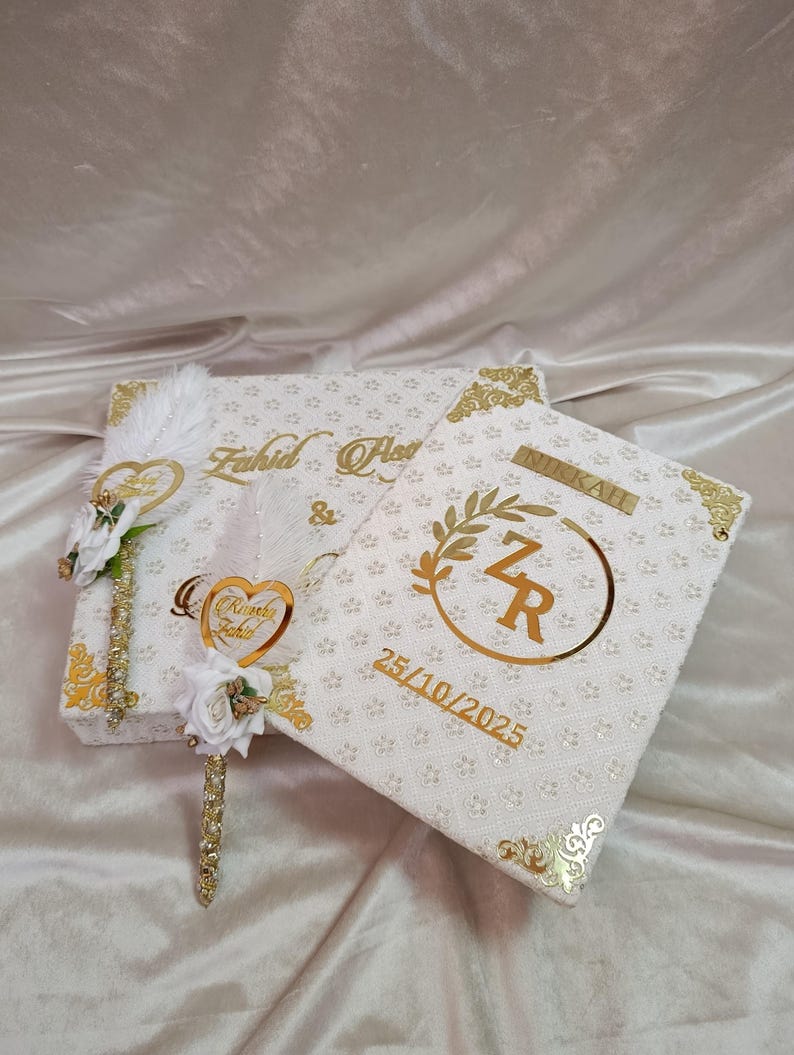 May include: Two white decorative boxes with gold detailing. One box features the names "Fahid & Asma" in gold script. The other box has the initials "ZR" within a gold wreath, with the date "25/10/2025" in gold.