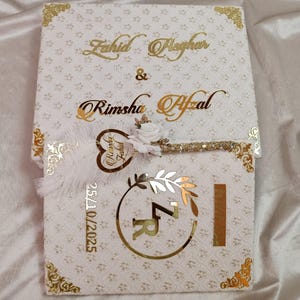 May include: A white wedding invitation box with gold detailing and floral patterns. The names "Zahid Pleghar" and "Rimsha Afzal" are in gold script. A key-shaped charm and the date "25/10/2025" are also visible.