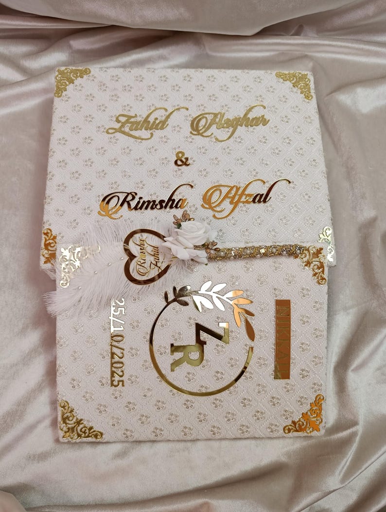 May include: A white wedding invitation box with gold detailing and floral patterns. The names "Zahid Pleghar" and "Rimsha Afzal" are in gold script. A key-shaped charm and the date "25/10/2025" are also visible.