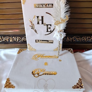 Puede incluir: Un juego de cajas de boda blancas con detalles dorados. El juego incluye una caja y un artículo en forma de libro, ambos con letras doradas y esquinas decorativas. El libro tiene la palabra "NIKKAH" y las iniciales "H & E". Se incluye un bolígrafo decorativo de plumas.