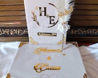 May include: A white wedding box set with gold detailing. The set includes a box and a book-shaped item, both with gold lettering and decorative corners. The book has the word "NIKKAH" and the initials "H & E". A decorative feather pen is included.