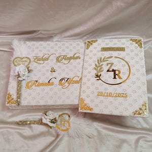 May include: A white wedding box set with gold detailing. The set features a box with names in gold script, a smaller book with the word "NIKKAH" and initials, and a decorative feather pen. The date 25/10/2025 is also visible.