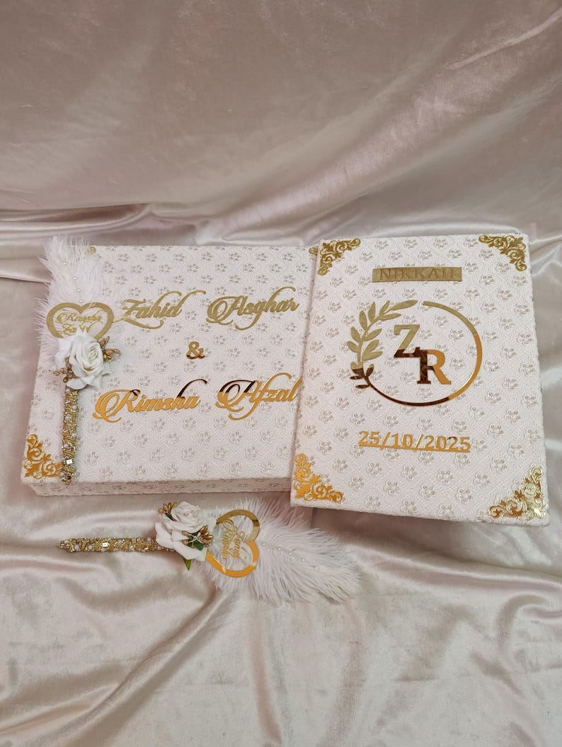 May include: A white wedding box set with gold detailing. The set features a box with names in gold script, a smaller book with the word "NIKKAH" and initials, and a decorative feather pen. The date 25/10/2025 is also visible.