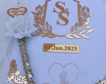 May include: A white wedding guest book with gold detailing. Features a gold emblem with the initials "SS", the date "11.Jan.2025", and decorative corner pieces. Includes a pen with a white rose and a heart-shaped charm.