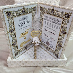 May include: A wedding gift box featuring two framed certificates with ornate gold and brown detailing on a white background. The box itself is white with a lace pattern. A gold heart ornament and white rose are in the center.